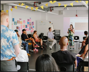 A woman stands and speaks in front of a seated group during a workshop or presentation in a  modern classroom. Attendees sit in a semi-circle, listening attentively. The room features a projector screen, and yellow bunting hanging from the ceiling.