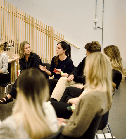 A woman is speaking in a group discussion, holding a colorful object. Others are seated around her, listening attentively in a casual, collaborative setting.