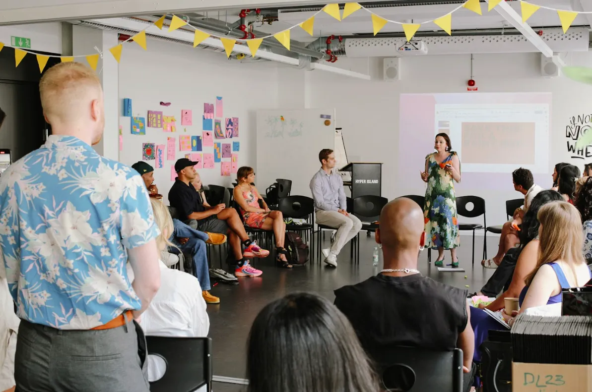 A speaker addresses a diverse group of adults in a bright room with yellow flags and art, during a casual presentation.