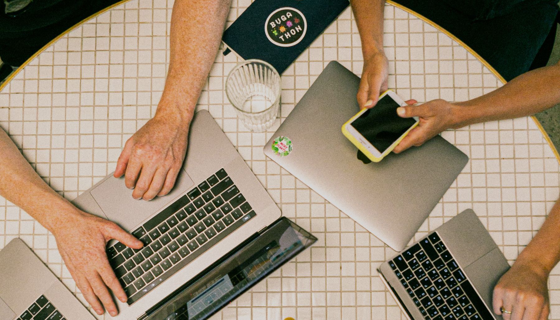 A group of people collaborating at a tech event, seen from above. They are using laptops and a smartphone on a modern, tiled table, suggesting teamwork and problem-solving.