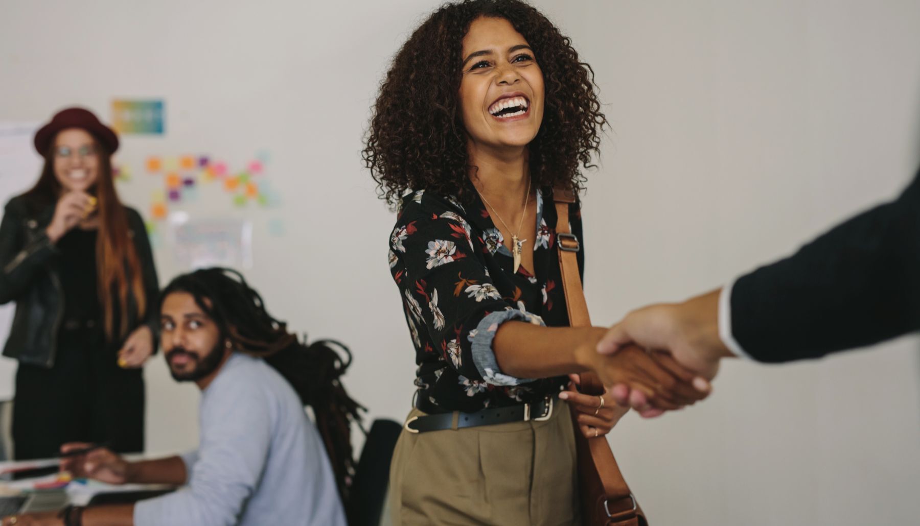 Close-up of a business development manager laughing happily as she shakes hands with a person. In the background, in a casual work environment, two colleagues watch the scene, smiling.