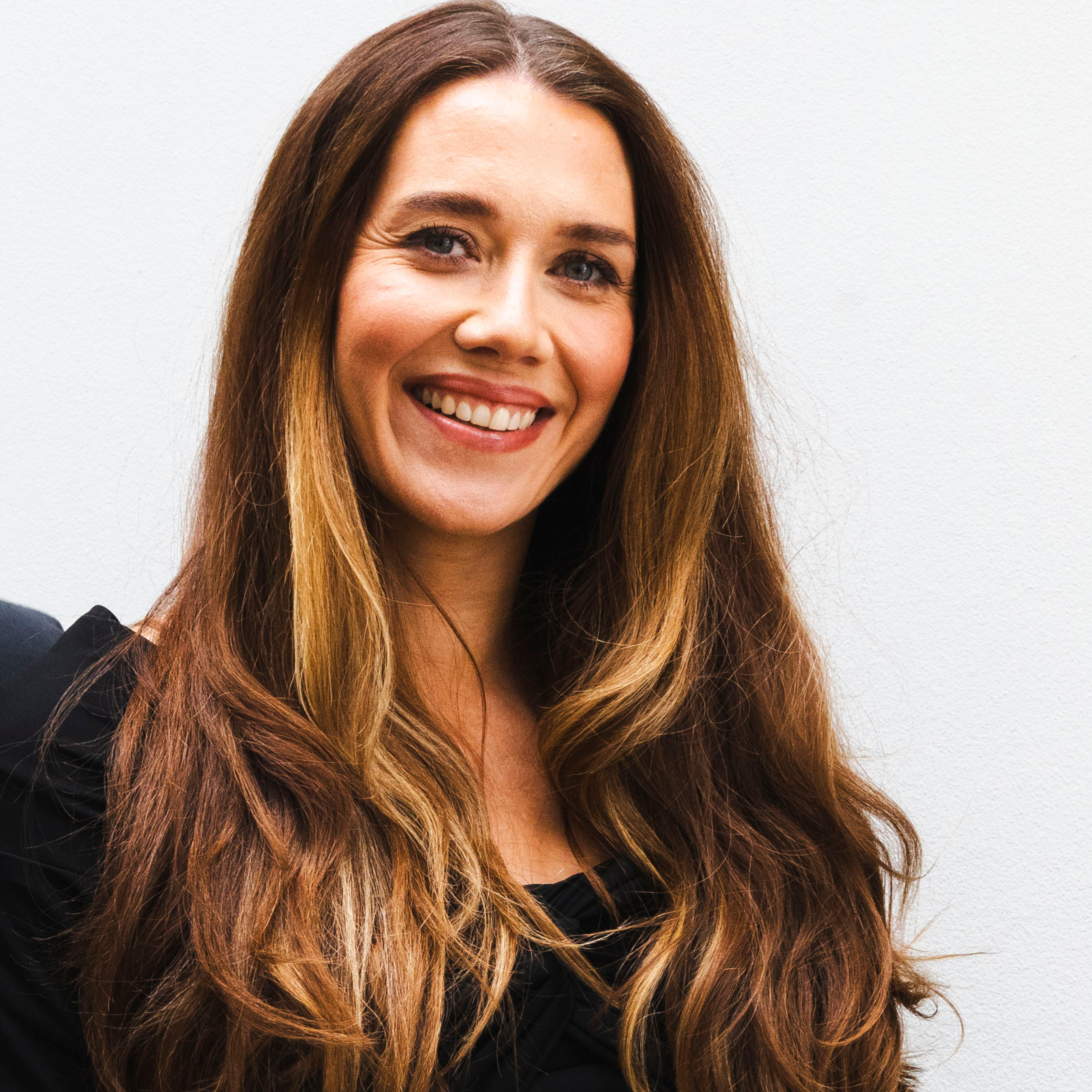 A smiling woman with long, flowing brown hair, streaked with lighter highlights, looks directly at the camera against a plain white background.