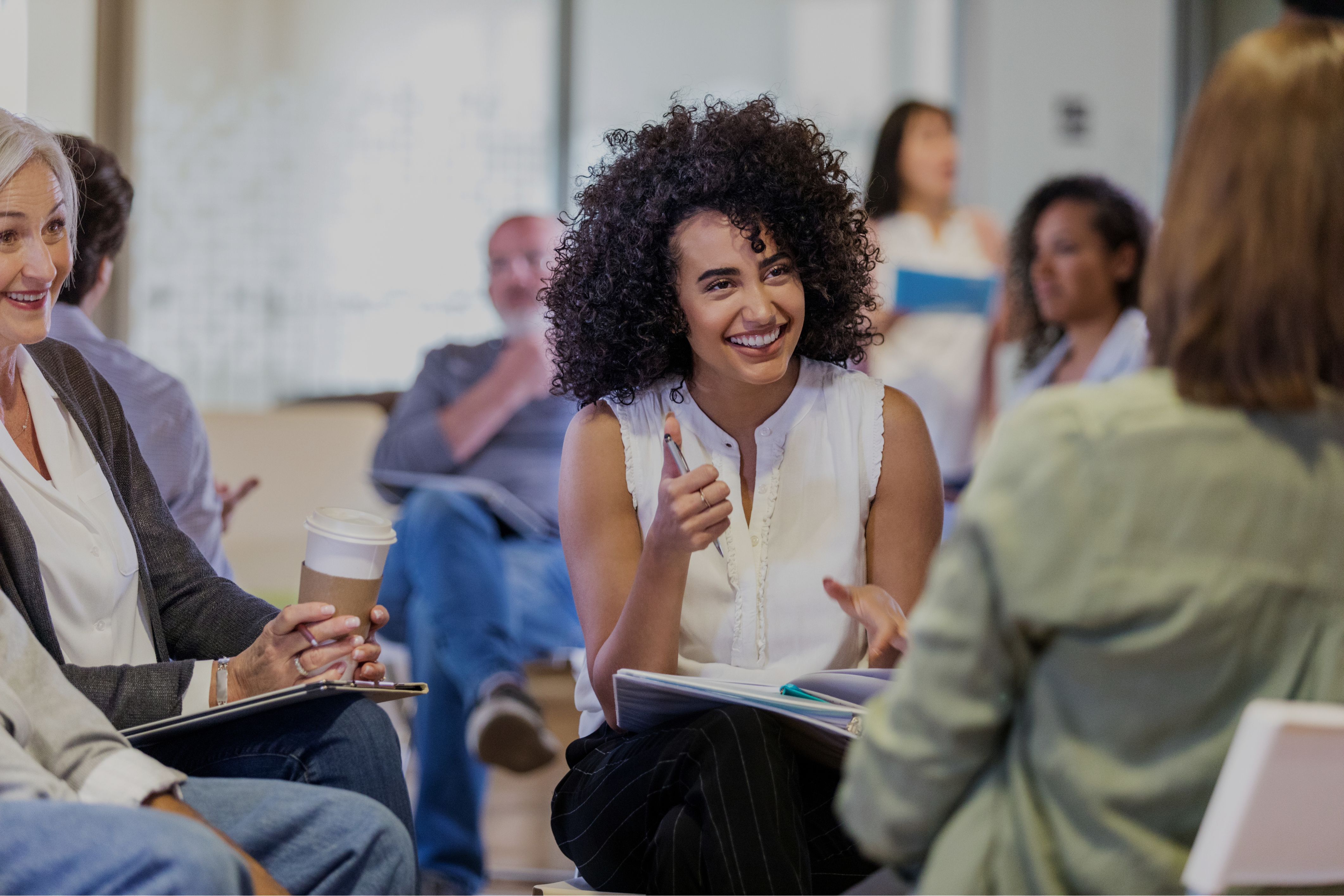 Woman holding a pen and a notebook, smiling while looking at a colleague in a room with other people, like a facilitation setting or a workspace.