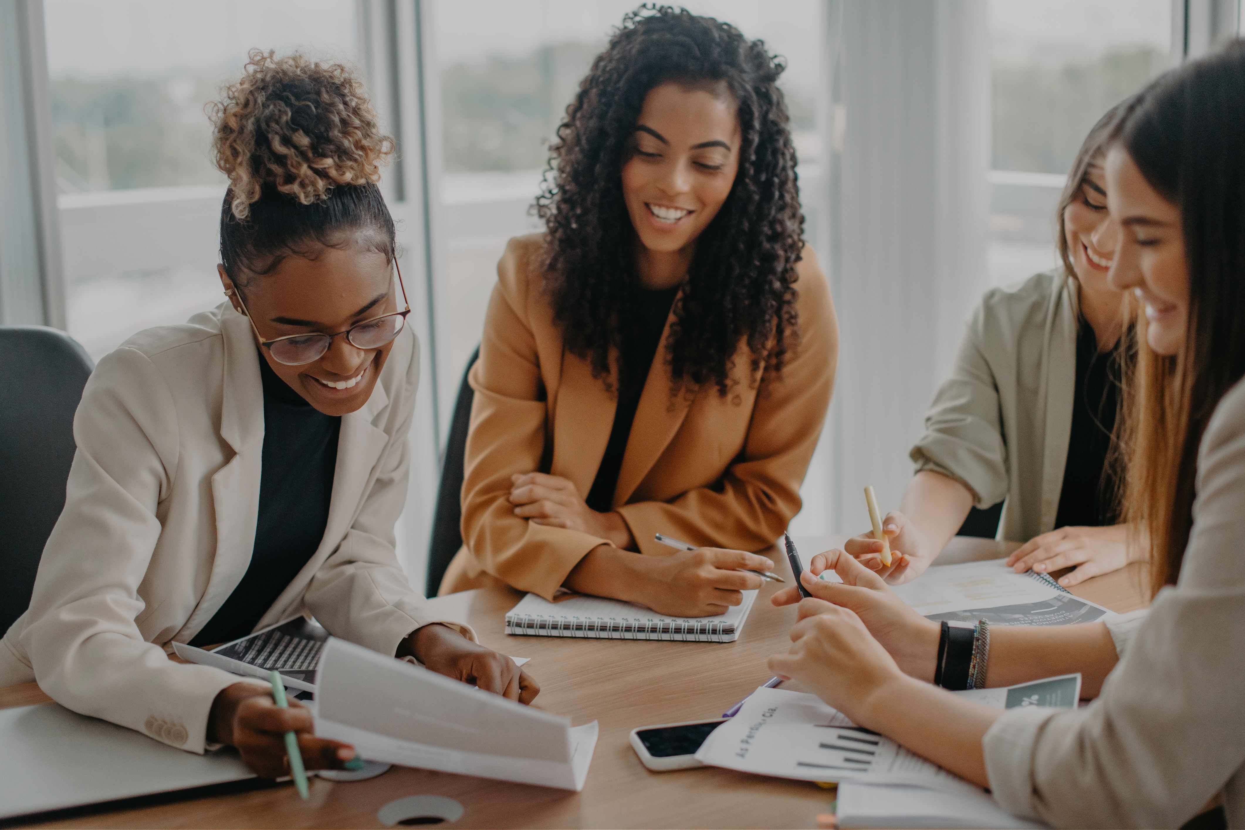 A group of female colleagues smiles and discusses a plan, analyzing data on papers and taking notes during a meeting in a modern office.