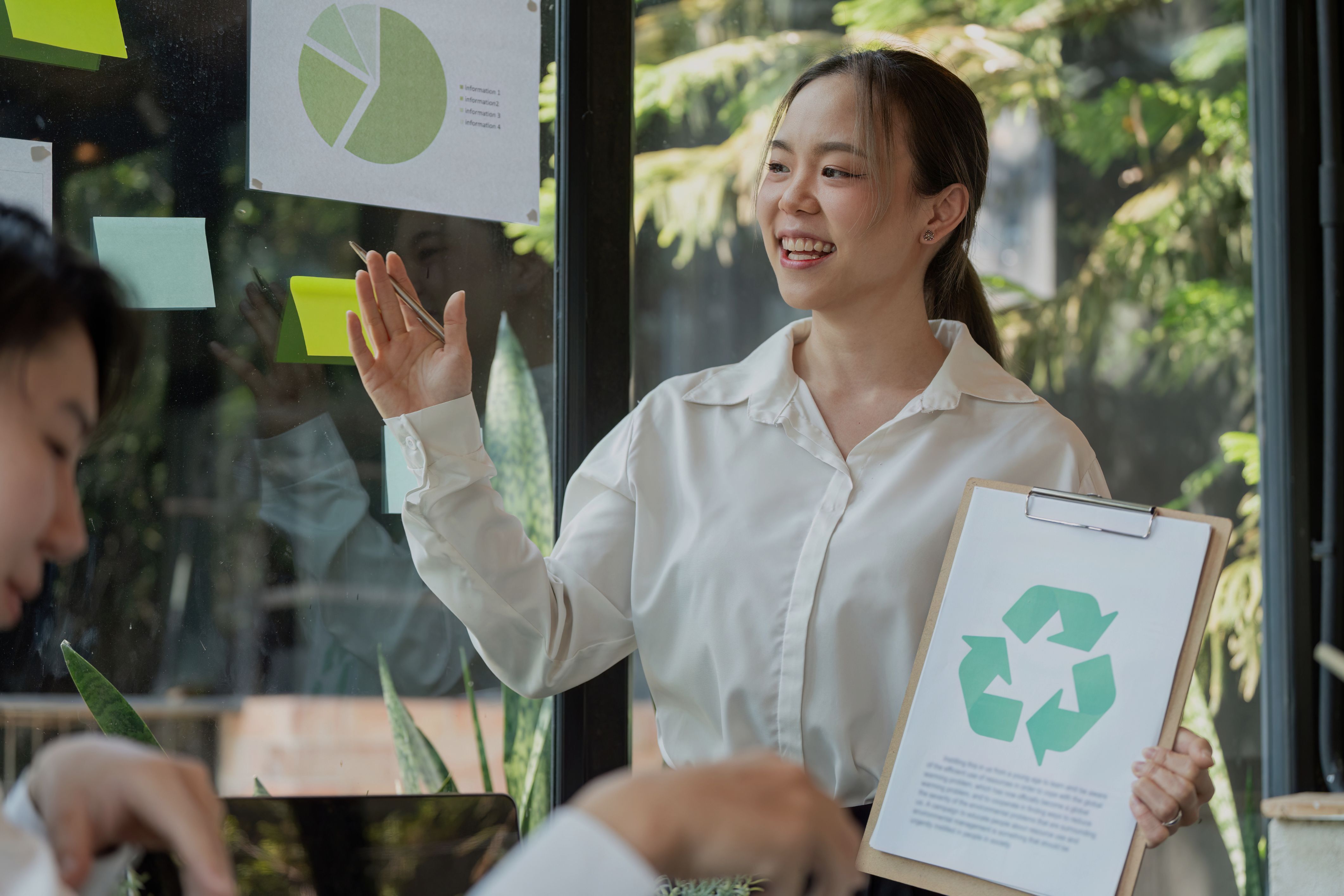 A manager presents a corporate sustainability strategy to a team, holding a clipboard with the recycling symbol and pointing to charts.