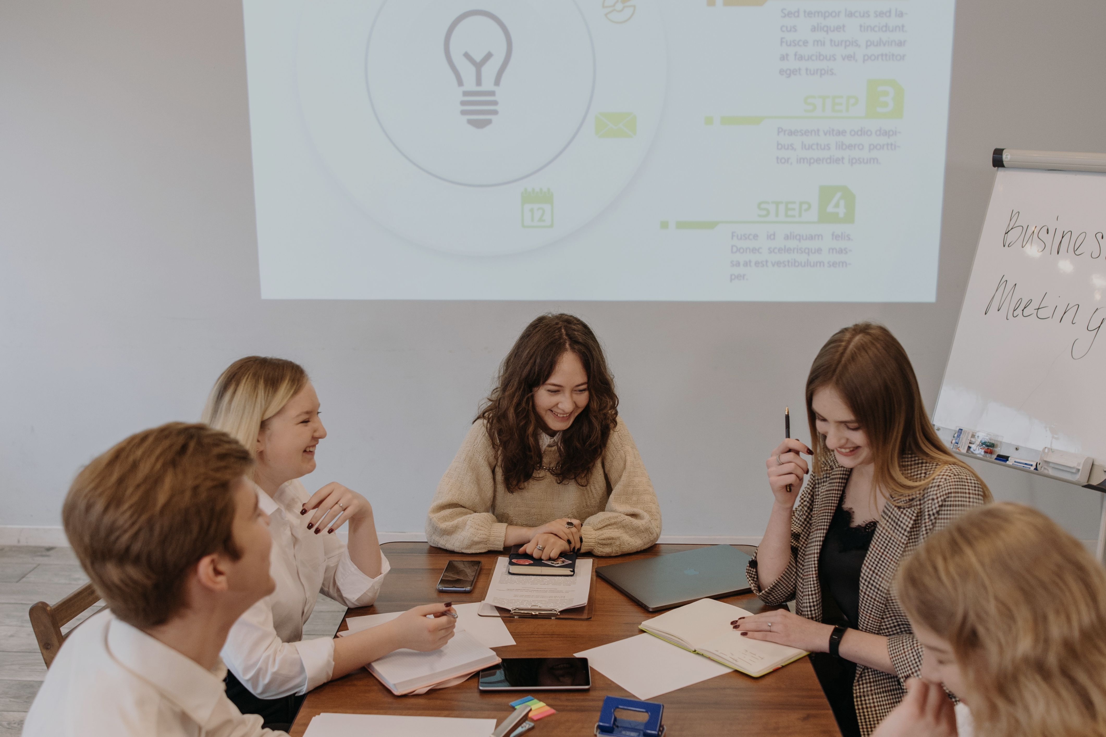 A smiling workgroup sits around a table during a meeting. In the background, a presentation shows an infographic with various steps, symbolizing the application of a strategy framework.