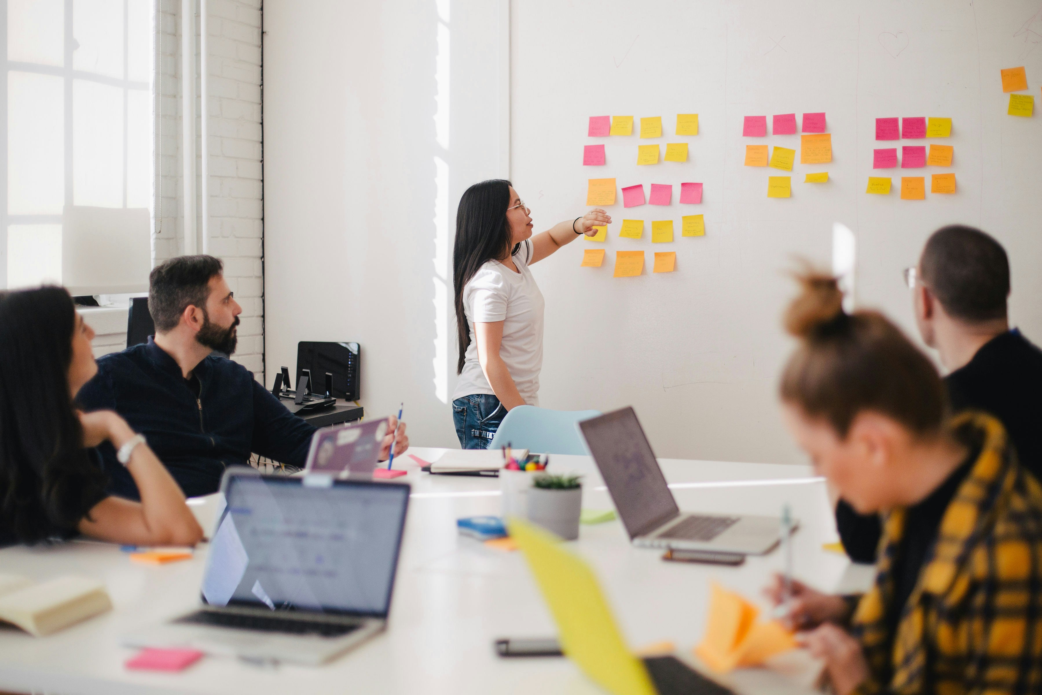 A woman leads a collaborative meeting in a bright, modern office, presenting ideas on a wall covered with colorful sticky notes to her attentive colleagues